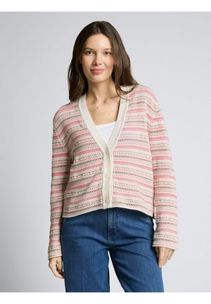 Kobieta o długich brązowych włosach, ubrana w biało-różowy pasiasty sweter szydełkowy na białym topie i niebieskich dżinsach, stojąca na szarym tle.