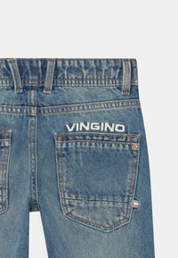 Takapuolelta näkyvät siniset farkut, joissa on ommeltu "VINGINO" -logo oikean takataskun yläpuolella ja pieni kolmivärinen kangastagi taskussa.