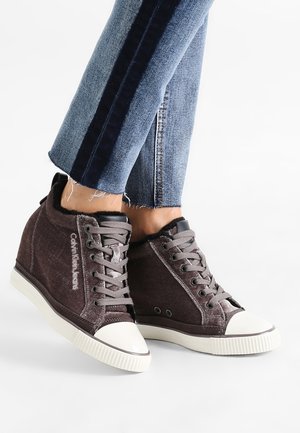 Zapatillas de caña alta de color marrón oscuro con punteras y suelas de goma blanca, cordones grises y el logotipo "Calvin Klein Jeans", usadas con jeans azules remangados.