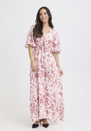 Vrouw die glimlacht, gekleed in een witte maxi-jurk met een roze bloemenprint, vlindermouwtjes en een taille met ceintuur, gecombineerd met zwarte hoge hakken.