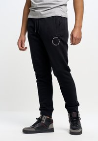 Schwarze Sweatpants aus einem Baumwollgemisch mit enganliegendem Design, elastischem Bund und weißem, rundem Logo am Oberschenkel. Kombiniert mit High-Top-Sneakern.