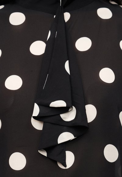 Μαύρη μπλούζα με μεγάλα λευκά πουά, με ρέον ζωνάκι στο λαιμό και απαλό, ελαφρύ ύφασμα. Ομαλή υφή με άνετη εφαρμογή.