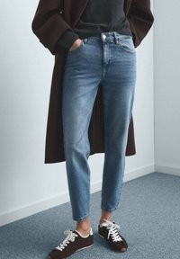 Jeans in denim azzurro chiaro con vestibilità affusolata, cinque tasche e lievi segni di usura. Abbinati a sneaker marrone scuro e un maglione nero.