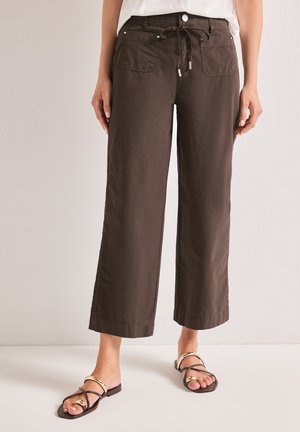 Femme portant un pantalon marron coupe large et courte, avec poches avant, taille nouée, et des sandales noires avec des détails dorés.