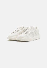 Bílé kožené tenisky s hladkým povrchem, s plochými tkaničkami, gumovou podrážkou a nenápadným brandingem na boku.