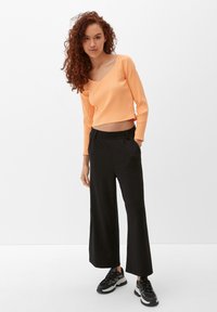Geribbelde, cropped, lange mouwen top in een levendig oranje; gecombineerd met losse zwarte broek. Zwarte sportschoenen maken de outfit compleet.
