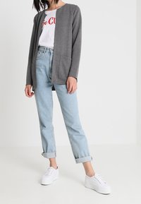 Szary długi kardigan z okrągłym dekoltem i przednimi kieszeniami, noszony na białej koszulce z czerwonym nadrukiem, zestawiony z jasnoniebieskimi podwiniętymi dżinsami i białymi tenisówkami.