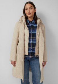 Beige lange jas met een afneembare capuchon over een crèmekleurige gewatteerde vest, gedragen over een blauwe geruite blouse, gecombineerd met blauwe jeans. Glad van textuur.