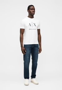 Λευκό βαμβακερό t-shirt με εκτυπωμένα τα "AX" και "ARMANI EXCHANGE" σε ναυτικό μπλε. Συνδυασμένο με σκούρα μπλε τζιν και λευκά αθλητικά παπούτσια, σε όρθια στάση.
