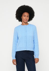 Gaiši zils adīts kardigans ar ietīta rakstu, apaļu kaklu, ribināt�ām aprocēm un piecu pogu aizdari, apvienots ar tumšiem biksēm.