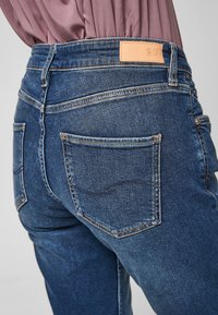 Tmavě modré džíny s klasickým střihem, vyznačující se světlou koženou nášivkou na pásku, dvěma zadními kapsami a kontrastním prošitím.