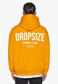 Gul hættejakke med stor hvid tekst "DROPSIZE" over "CRIME CLUB" og mindre tekst nedenfor. Har en kengurulomme og justerbar hætte.