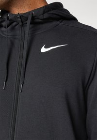 Sudadera con cremallera negra de tejido texturizado. Cuenta con capucha ajustable con cordón y un destacado logo blanco de Nike en el pecho.