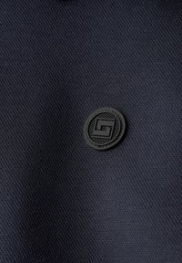 Tissu noir texturé avec un petit patch rond noir présentant un logo carré stylisé en son centre.
