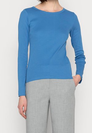 Person, die ein eng anliegendes blaues Langarmoberteil und hellgraue Hosen trägt, mit einer Hand in der Tasche vor einem schlichten Hintergrund.