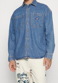 Camicia in denim blu con due tasche sul petto, bottoni dorati e orlo arrotondato. Il tessuto appare morbido con una texture leggermente sbiadita.