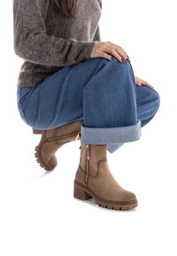 Stivaletti in suede marrone con zip laterale, tacco a blocco e suola texturizzata, abbinati a pantaloni blu con risvolto e un maglione grigio.
