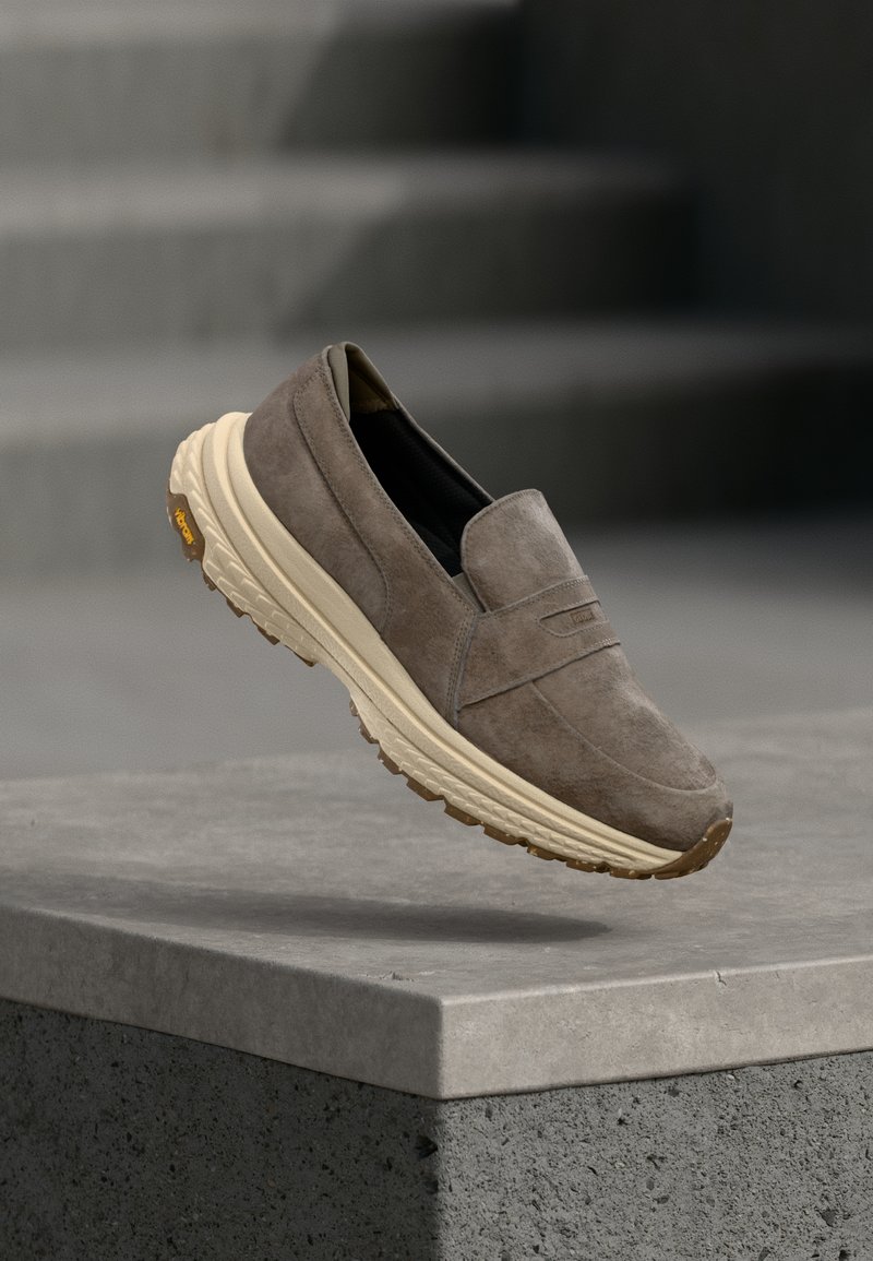 Chaussure en daim marron sans lacets avec une semelle épaisse crème flottant au-dessus d'un rebord en béton gris, avec des marches floues en arrière-plan.