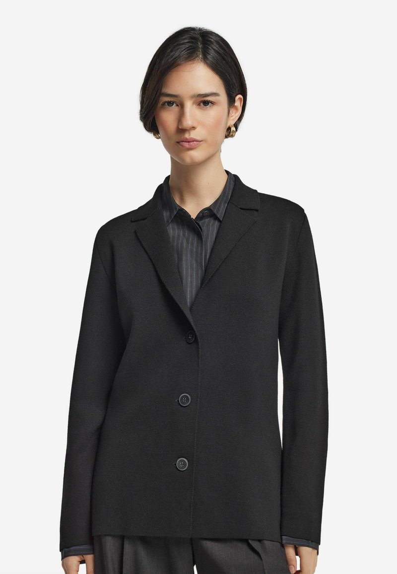 Schwarzer Blazer aus strukturiertem Material mit Reverskragen, dreireihiger Knopfleiste und langen Ärmeln. Über einem gestreiften Hemd getragen.