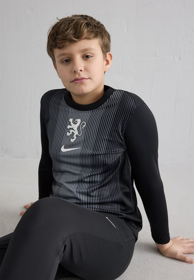 Băiat tânăr stând pe podea, purtând un tricou sport negru cu mâneci lungi, cu emblemă de leu și logo Nike, pantaloni sport negri, fundal neutru.