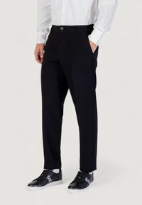 Pantaloni neri su misura con una texture liscia, dotati di tasche laterali e chiusura con bottone, abbinati a sneakers nere.