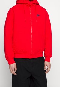 Person som bär en klar röd Nike zip-up hoodie med huva och dragskor, i kombination med löst sittande svarta byxor.