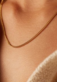 Dunne gouden ketting ligt op een gladde huidoppervlak, met een eenvoudige gedraaide design en een gepolijste afwerking. Zacht beige stof is zichtbaar in de nabijheid.