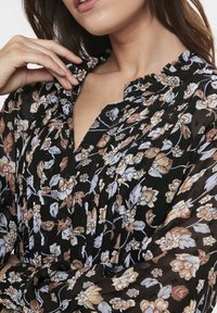 Blusa negra con flores y cuello con volantes, que presenta un patrón de flores en beige y azul, cierre de botones y textura de tela suave y translúcida.