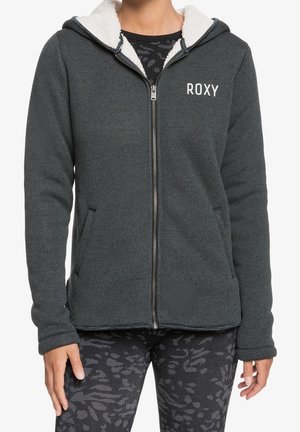 Vrouw draagt een donkergrijze ritssluiting met capuchon jas met witte fleece voering en het logo "ROXY", gecombineerd met zwarte legging met patroon.