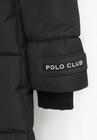 Svart pufferjacka ärm med en tygpatch där det står "POLO CLUB" i vit text. Slät textur med ribbad svart mudd.
