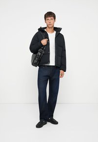 Sort pufferjakke med lynlås og trykknaplukning, båret over en hvid t-shirt og mørke denimjeans, med sorte sko og en skuldertaske.
