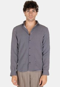 Giovane con capelli ricci che indossa una camicia a bottoni con un motivo geometrico in bianco e nero e pantaloni beige, in piedi davanti a uno sfondo bianco.