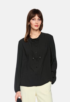 Woman with brown hair wearing a black button-up blouse with tie accents and cream trousers, holding a black handbag against a plain background.