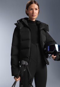 Žena v černé lyžařské bundě a kalhotách drží lyžařské hole a černou lyžařskou helmu s reflexními brýlemi na šedém pozadí.