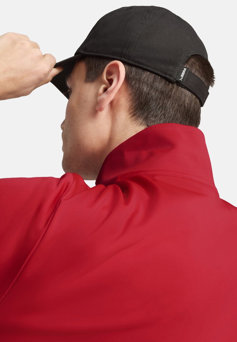 Veste de sport rouge avec un col haut et une casquette noire. La veste est en tissu lisse, tandis que la casquette a un design structuré et une visière plate.