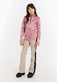 Lichtroze top met lange mouwen, een opstaande kraag en een halve ritssluiting. Gecombineerd met beige broeken met zwarte zijstrepen en zwarte laarzen.