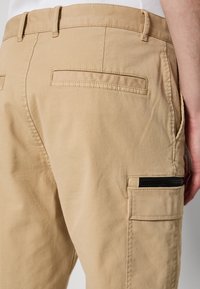 Pantalones cargo en color caqui con una tela texturizada, que cuentan con bolsillos traseros, un bolsillo lateral con cremallera y presillas para cinturón.