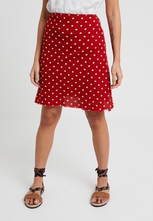 Mujer con falda roja con lunares blancos, blusa de encaje blanca y sandalias marrones con tiras en el tobillo, de pie contra un fondo liso.