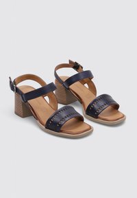 Sandalias de tacón con correas de cuero azul marino oscuro, suela beige y un tacón de bloque de madera texturizado. Presenta patrones recortados en las correas.
