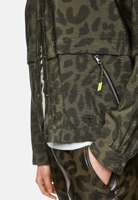 Khaki leopard print jacket, lightweight fabric, snap closure, zippered pocket with neon accent, ribbed cuffs, layered over a white shirt.
