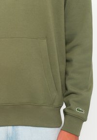 Olijfgroene hoodie met een kangoeroezak, ribgebreide manchetten en zoom. Heeft een klein geborduurd krokodillenlogo op de mouw. Van een katoenmix.