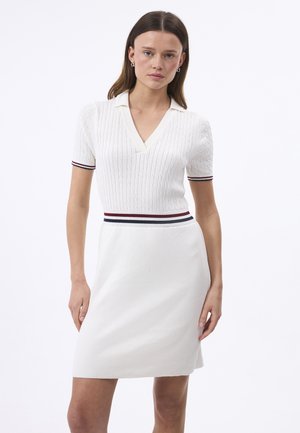 Kvinde iført en hvid kortærmet strikket kjole med krave og rød, hvid og blå stribet kant på ærmer og talje.