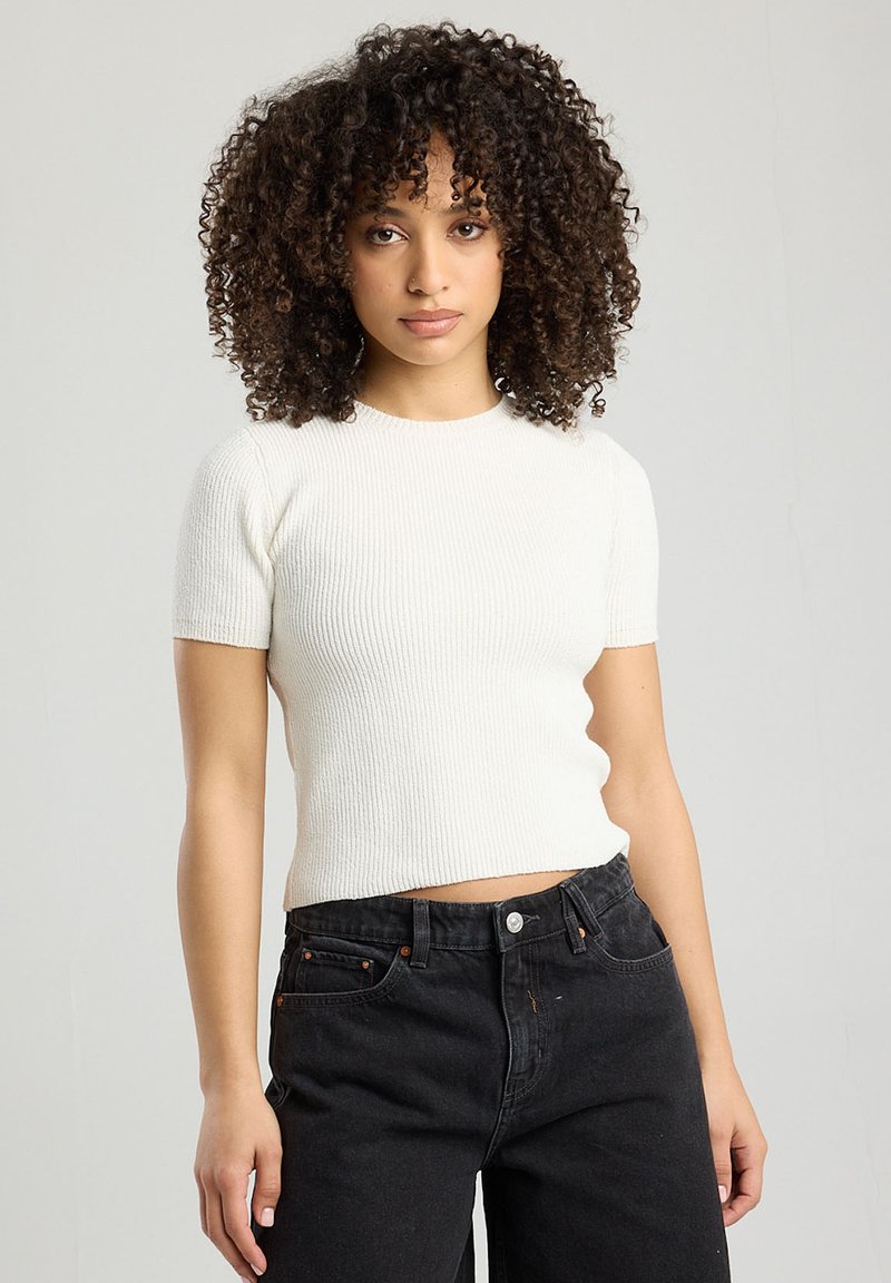 White ribbed short-sleeve top with a round neckline, fitted shape, and textured fabric, paired with high-waisted black jeans.