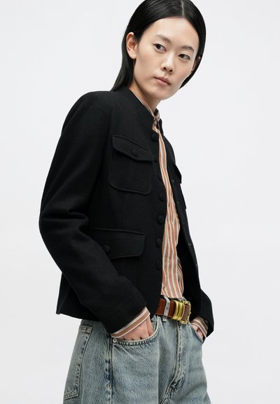 Sort cropped jakke med knapper foran, to brystlommer, kombineret med en stribet skjorte og blå jeans. Glat tekstur, tætsiddende design.