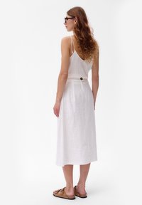 Witte linnen jurk met een licht uitlopende silhouette, voorzien van een knoopdetail op de rug en dunne schouderbanden. Draag met bruine sandalen.