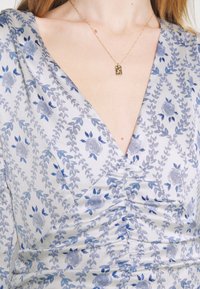 Femme portant une robe florale gris argenté avec un détail froncé et un collier pendentif rectangulaire doré sur une chaîne fine.