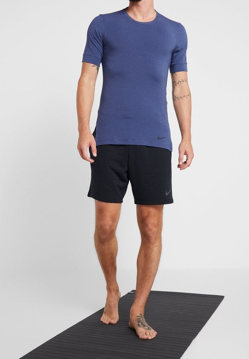 Lyhyttä hihaista tummansinisestä urheiluvaatetuksesta valmistettua paitaa, joka on yhdistetty mustiin shortseihin; Nike-logo paidassa ja shortseissa; matto alla.