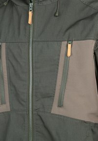 Grön och brun zip-up-jacka med två framfickor, en texturerad yta, dragsko i huvan och kontrasterande tygpaneler.