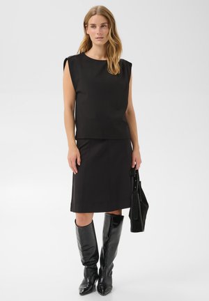 Femme portant un haut noir sans manches, une jupe noire mi-longue, des bottes noires montantes, tenant un sac à main noir, debout sur un fond uni.