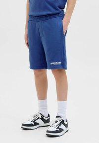 Blå idrottsshorts med resårmidja, sidofickor och en vit logotyp vid fållen. Bärs med vita strumpor och svart-vita sneakers.